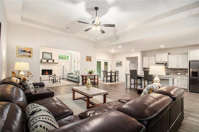 living room with a ceiling fan, wood finished floors, recessed lighting, a tray ceiling, and a brick fireplace