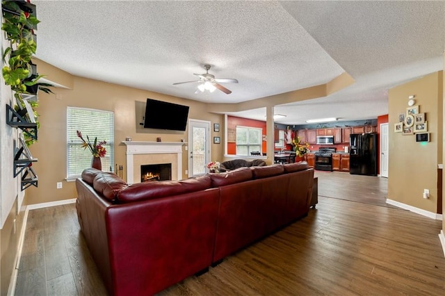 living area featuring dark wood-type flooring, a warm lit fireplace, ceiling fan, and baseboards