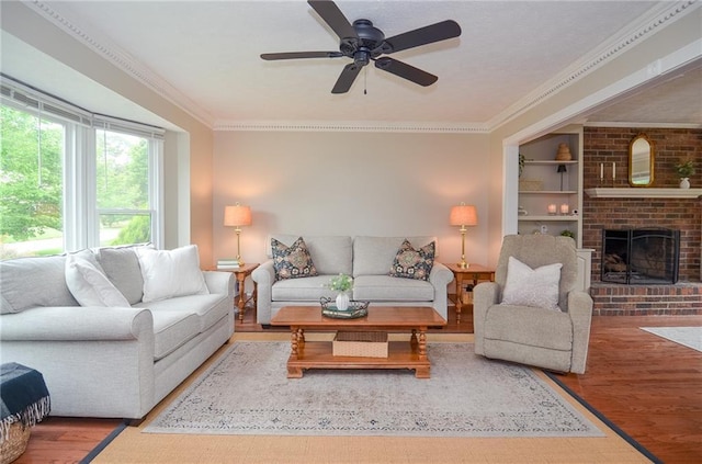 living room with ceiling fan, crown molding, a brick fireplace, built in shelves, and wood finished floors