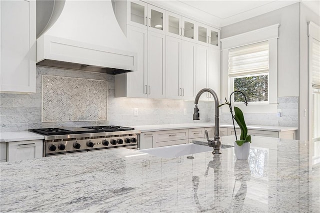 kitchen with custom exhaust hood, backsplash, glass insert cabinets, light stone countertops, and range