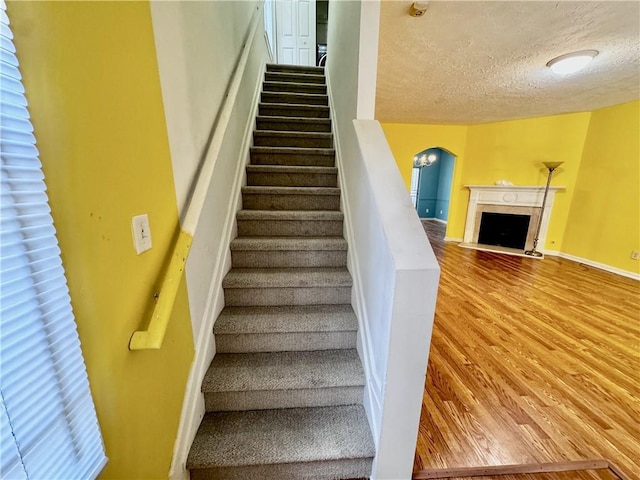 stairs featuring wood finished floors, a fireplace with flush hearth, a textured ceiling, baseboards, and arched walkways