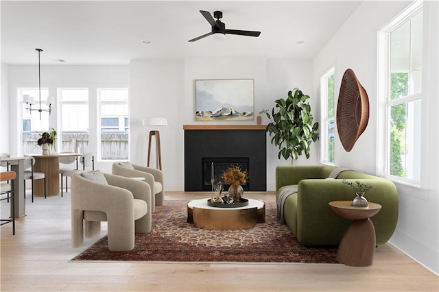 living area featuring wood finished floors, a glass covered fireplace, ceiling fan with notable chandelier, and a wealth of natural light