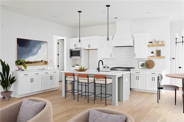 kitchen with open shelves, premium range hood, stainless steel microwave, white cabinets, and a sink