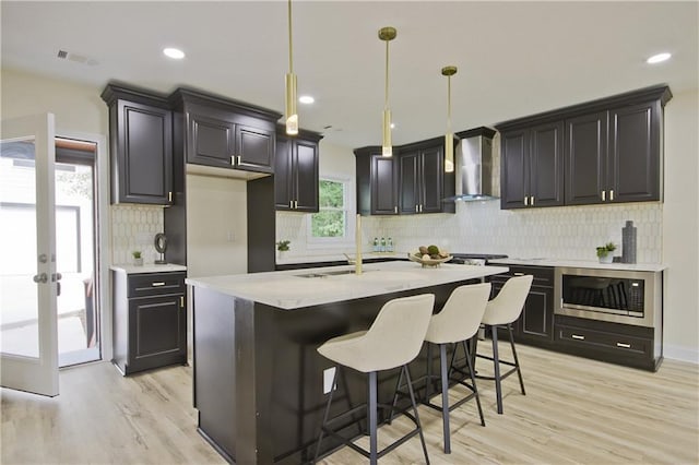 kitchen featuring stainless steel microwave, wall chimney exhaust hood, pendant lighting, and light hardwood / wood-style flooring