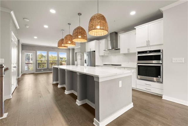 kitchen with stainless steel appliances, a kitchen island with sink, wall chimney range hood, decorative light fixtures, and dark hardwood / wood-style floors