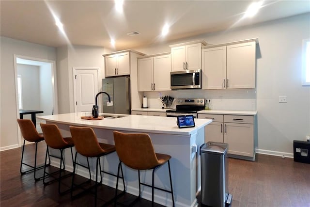 kitchen with sink, a center island with sink, appliances with stainless steel finishes, a kitchen bar, and dark hardwood / wood-style flooring