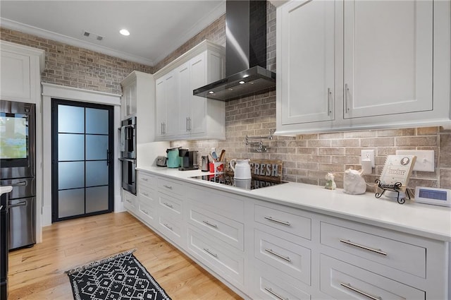 kitchen with double oven, white cabinetry, ornamental molding, wall chimney range hood, and light hardwood / wood-style flooring