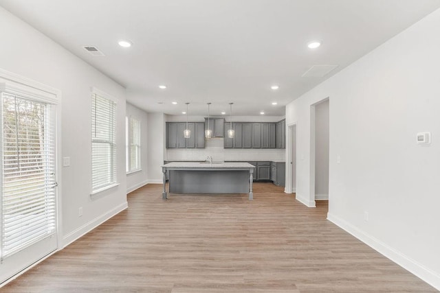 kitchen featuring gray cabinetry, a kitchen island with sink, wall chimney range hood, light wood-type flooring, and decorative light fixtures