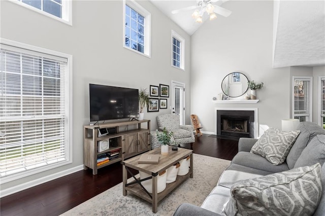 living room featuring baseboards, a fireplace with flush hearth, wood finished floors, high vaulted ceiling, and a ceiling fan