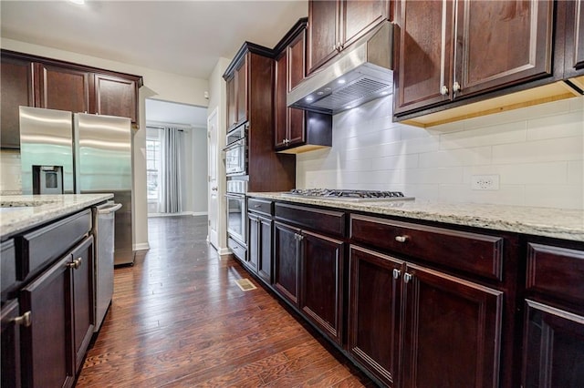 kitchen with stainless steel appliances, light stone counters, under cabinet range hood, decorative backsplash, and dark wood-style flooring