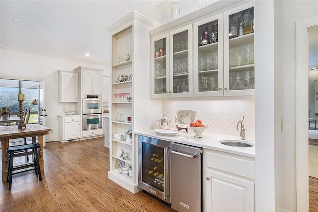 bar featuring beverage cooler, a sink, wood finished floors, decorative backsplash, and stainless steel double oven