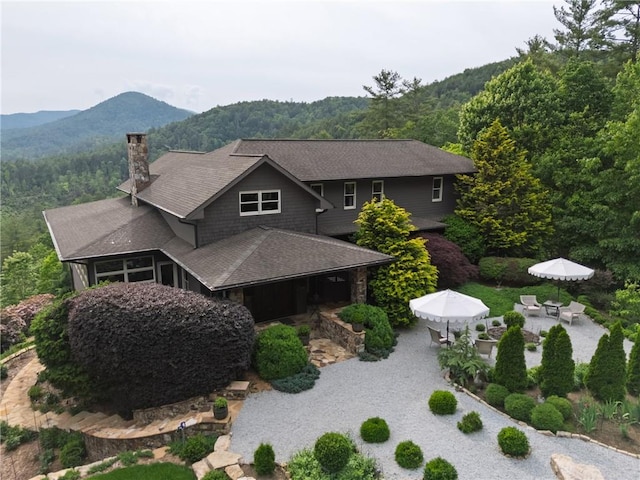 view of front facade with a mountain view, a chimney, a patio area, a wooded view, and outdoor dining space
