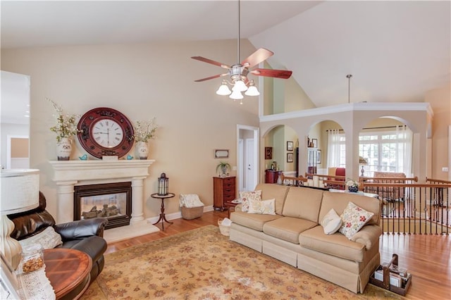 living area with ceiling fan, high vaulted ceiling, light wood-style flooring, baseboards, and a glass covered fireplace