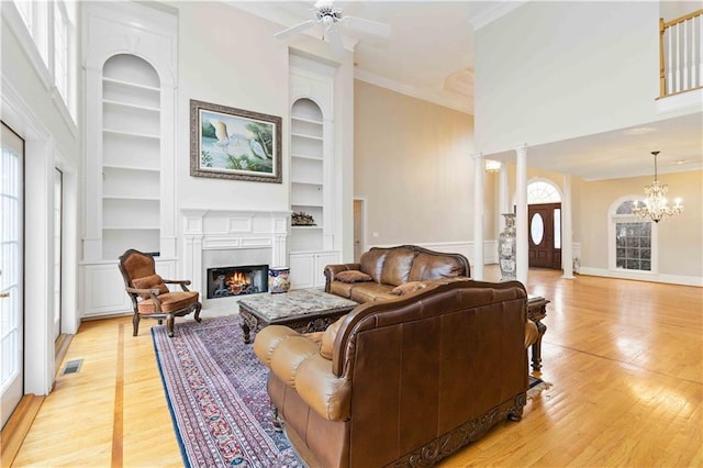 living room featuring built in features, light wood-type flooring, ceiling fan with notable chandelier, and ornate columns