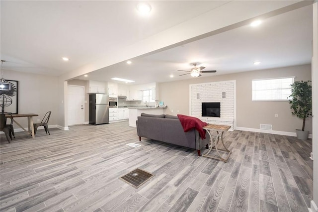 living room featuring baseboards, visible vents, ceiling fan, light wood-type flooring, and a brick fireplace