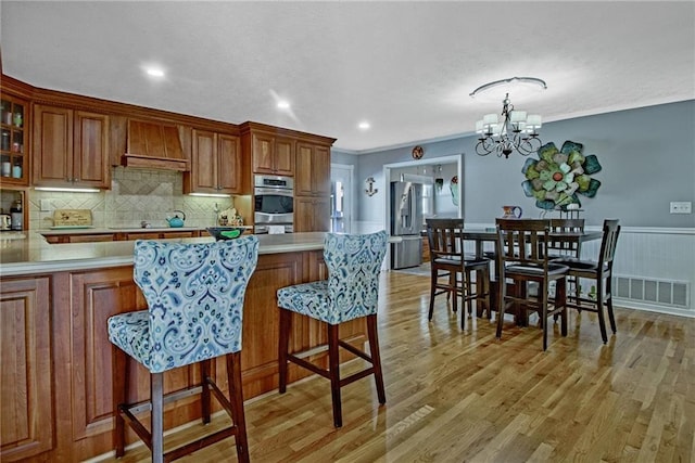 kitchen with hanging light fixtures, custom range hood, appliances with stainless steel finishes, light wood-type flooring, and a breakfast bar area