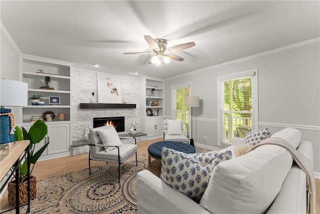 living room with built in features, a stone fireplace, a ceiling fan, crown molding, and light wood-style flooring