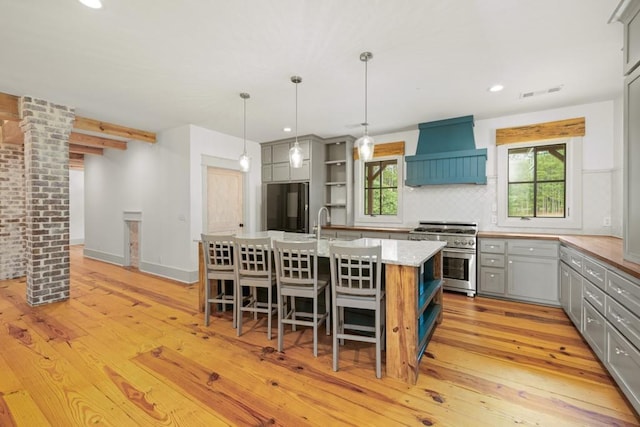 kitchen featuring custom exhaust hood, refrigerator, stainless steel range, an island with sink, and pendant lighting