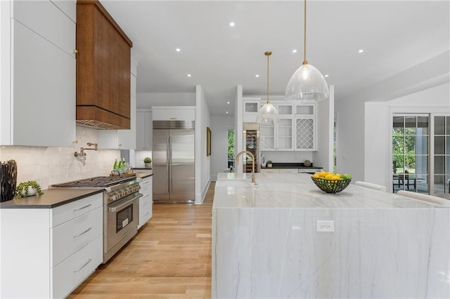 kitchen featuring light wood-type flooring, high end appliances, pendant lighting, white cabinets, and an island with sink