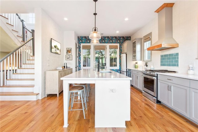 kitchen with sink, wall chimney range hood, pendant lighting, high end stainless steel range, and a center island with sink