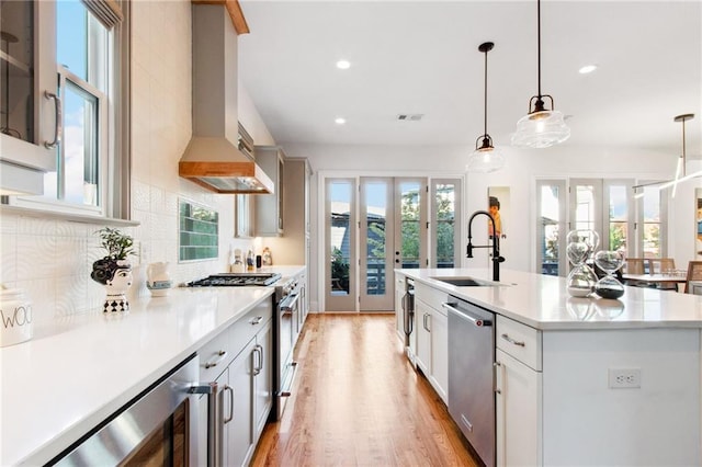 kitchen with sink, wall chimney exhaust hood, stainless steel appliances, decorative light fixtures, and a kitchen island with sink