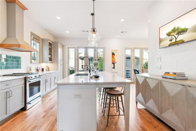 kitchen featuring wall chimney exhaust hood, high end stove, decorative light fixtures, a center island with sink, and gray cabinets