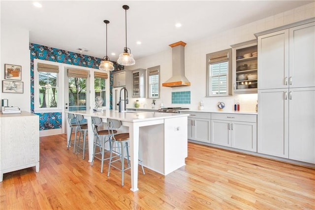 kitchen featuring gray cabinetry, wall chimney range hood, an island with sink, and hanging light fixtures