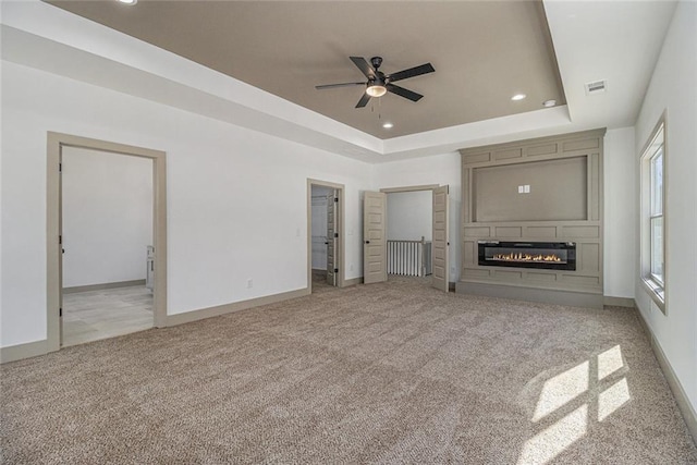 unfurnished living room with a raised ceiling, a large fireplace, light colored carpet, and ceiling fan