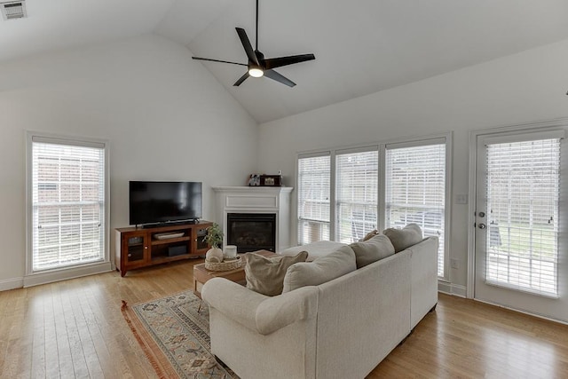 living room featuring high vaulted ceiling, light wood-style flooring, visible vents, a ceiling fan, and a glass covered fireplace