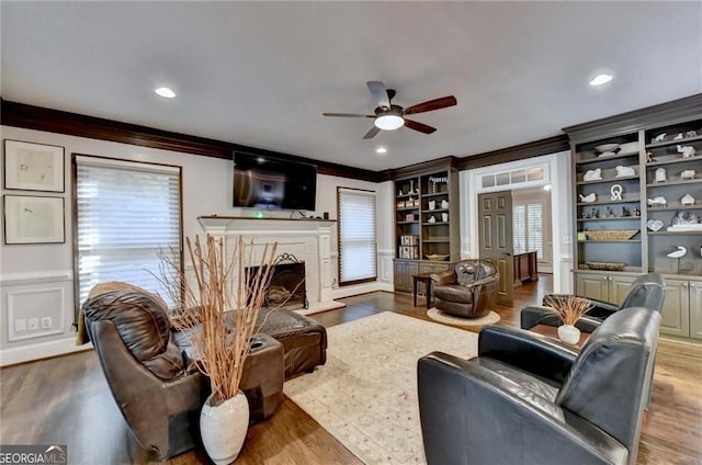 living area with wood finished floors, a ceiling fan, crown molding, and a fireplace with raised hearth