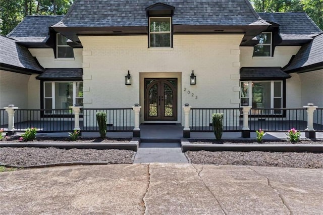 doorway to property featuring brick siding, covered porch, french doors, and roof with shingles