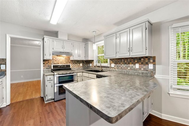 kitchen featuring double oven range, under cabinet range hood, a sink, a peninsula, and baseboards