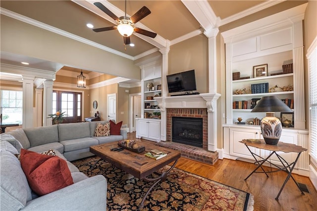 living area featuring a ceiling fan, decorative columns, crown molding, a brick fireplace, and wood finished floors