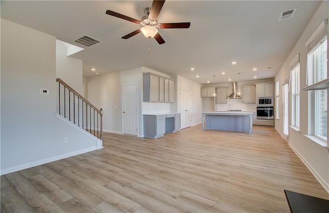 kitchen with gray cabinets, a kitchen island with sink, wall chimney exhaust hood, stainless steel appliances, and light hardwood / wood-style flooring