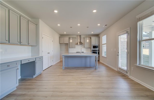 kitchen with decorative light fixtures, built in microwave, oven, wall chimney range hood, and a center island with sink