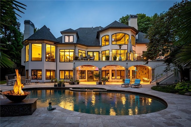 back house at dusk with a swimming pool with hot tub, a balcony, a patio, and an outdoor fire pit