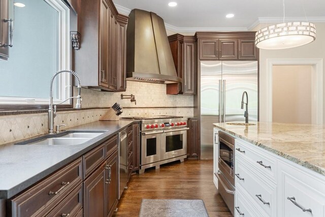 kitchen with sink, built in appliances, wall chimney exhaust hood, ornamental molding, and white cabinetry