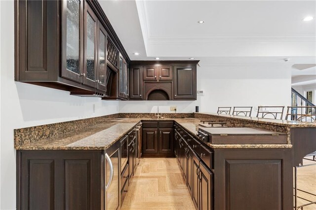 kitchen with a kitchen breakfast bar, ornamental molding, dark brown cabinetry, dark stone countertops, and light parquet flooring