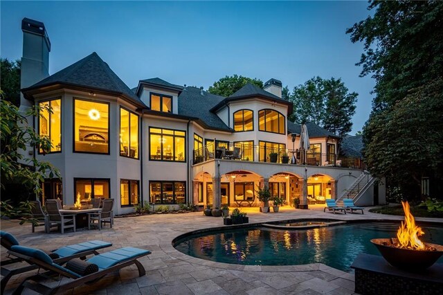 back house at dusk featuring a swimming pool with hot tub, a patio, a balcony, and an outdoor fire pit