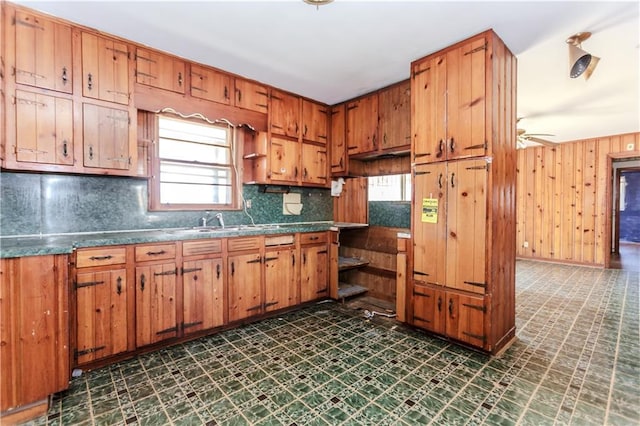 kitchen with dark floors, brown cabinets, backsplash, a ceiling fan, and wooden walls