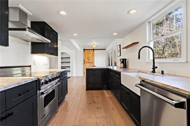 kitchen featuring sink, light hardwood / wood-style floors, stainless steel appliances, a barn door, and wall chimney exhaust hood