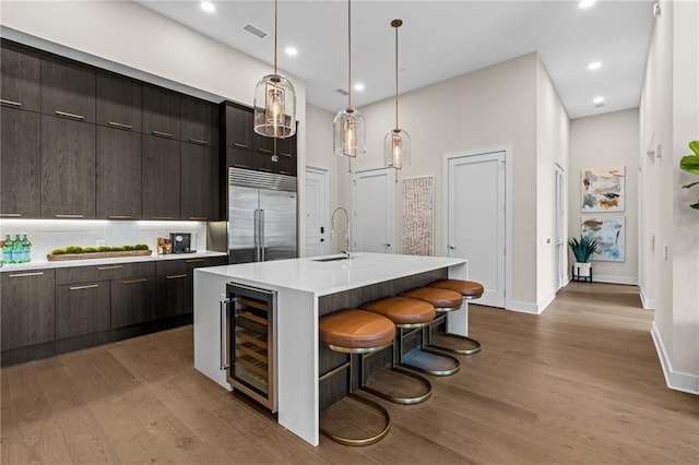 kitchen featuring beverage cooler, stainless steel built in refrigerator, light wood-type flooring, a center island with sink, and a breakfast bar area