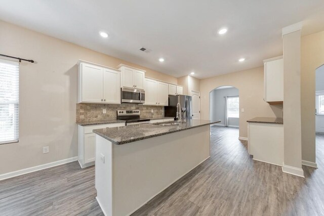 kitchen with appliances with stainless steel finishes, arched walkways, a center island with sink, light wood finished floors, and recessed lighting