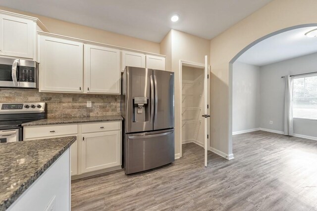 kitchen featuring stainless steel appliances, arched walkways, decorative backsplash, dark stone counters, and light wood-style floors
