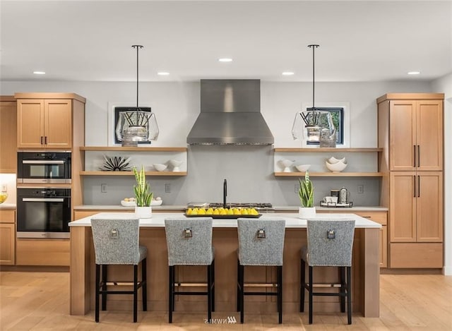 kitchen featuring wall chimney range hood, light hardwood / wood-style flooring, a breakfast bar, a center island with sink, and decorative light fixtures