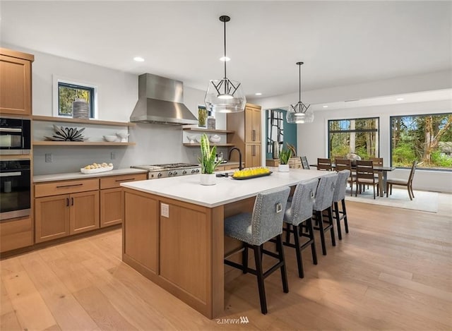 kitchen featuring wall chimney exhaust hood, stainless steel appliances, light hardwood / wood-style floors, and a center island with sink