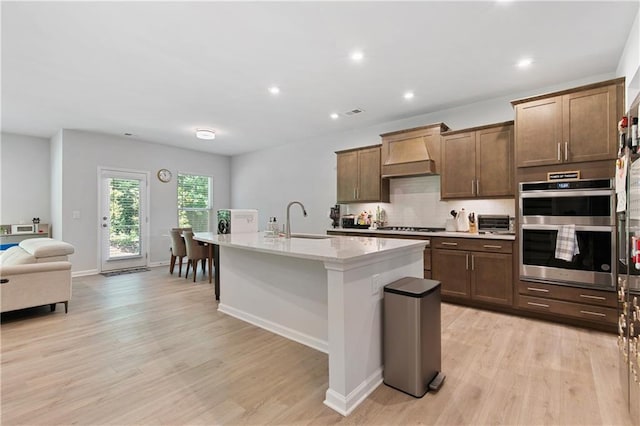 kitchen with light hardwood / wood-style flooring, stainless steel double oven, premium range hood, and a kitchen island with sink