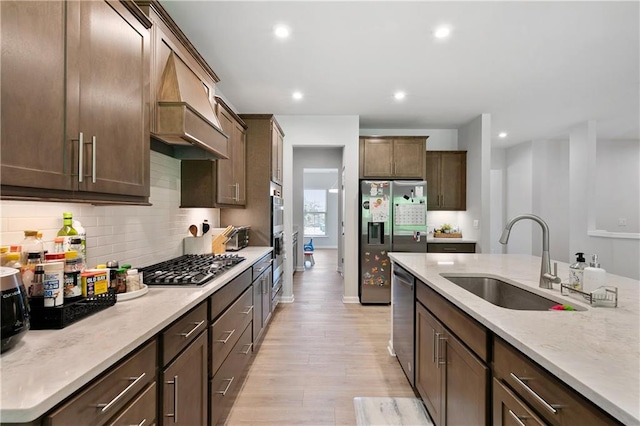 kitchen with stainless steel appliances, sink, custom range hood, dark brown cabinets, and light hardwood / wood-style flooring