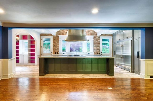 bar area with appliances with stainless steel finishes, wall chimney exhaust hood, light wood finished floors, built in features, and a decorative wall