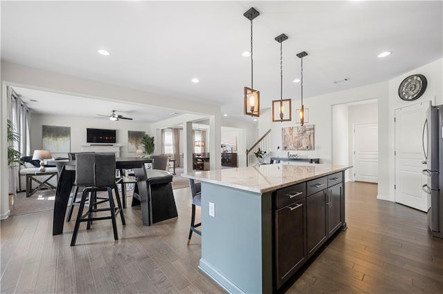 kitchen with pendant lighting, dark brown cabinets, a center island, light stone countertops, and ceiling fan